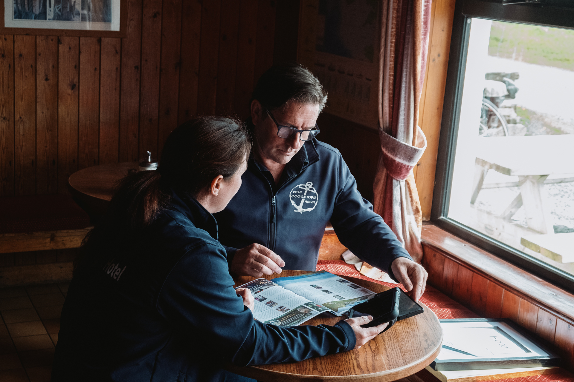 A man and a woman sit at a round table looking at a brochure and a computer tablet. 