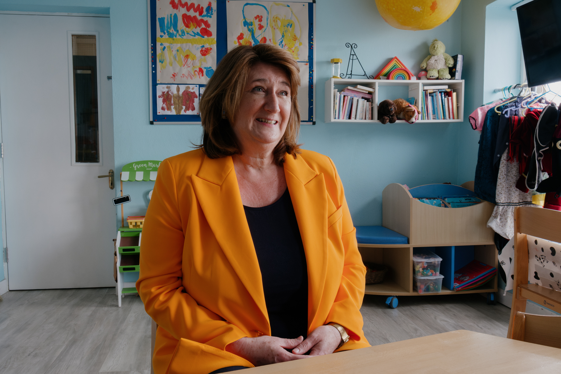 A woman sits in a classroom painted in bright colors wearing a bright orange jacket. 