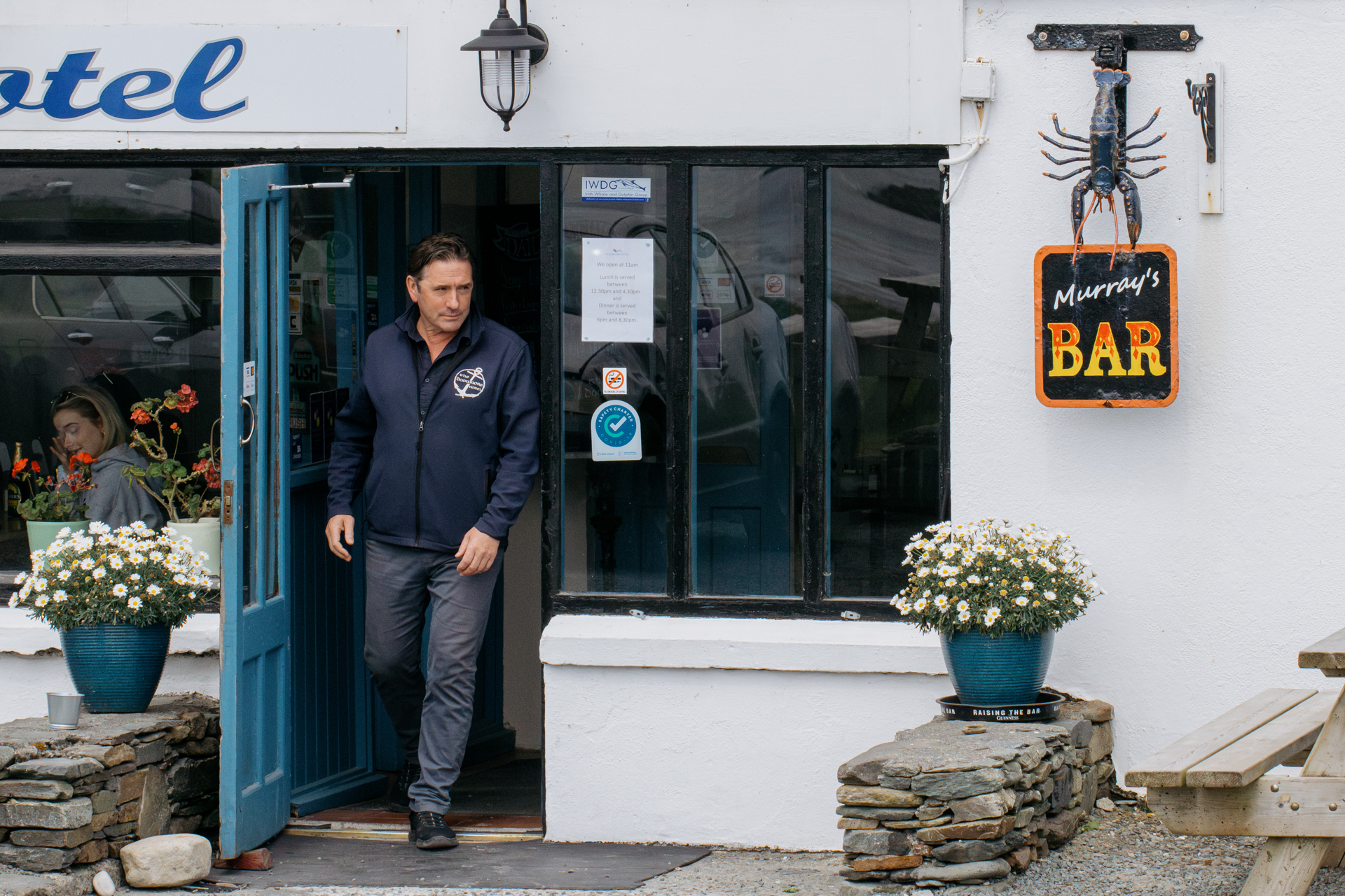 A man in a blue windbreaker exits a door of a white building. To the right, a sign says “Murray’s Bar” under a sculpture of a blue lobster. 