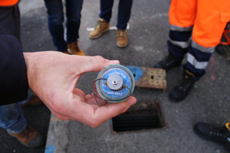 A closeup of a hand holding a blue FIDO sensor, with an elastic band for attaching it to pipes.