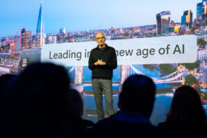 Satya Nadella is standing onstage with a presentation behind him. The image is a landscape of London and the words read 'Leading in the new age of AI'. There are silhouettes of audience members as they watch Satya present.