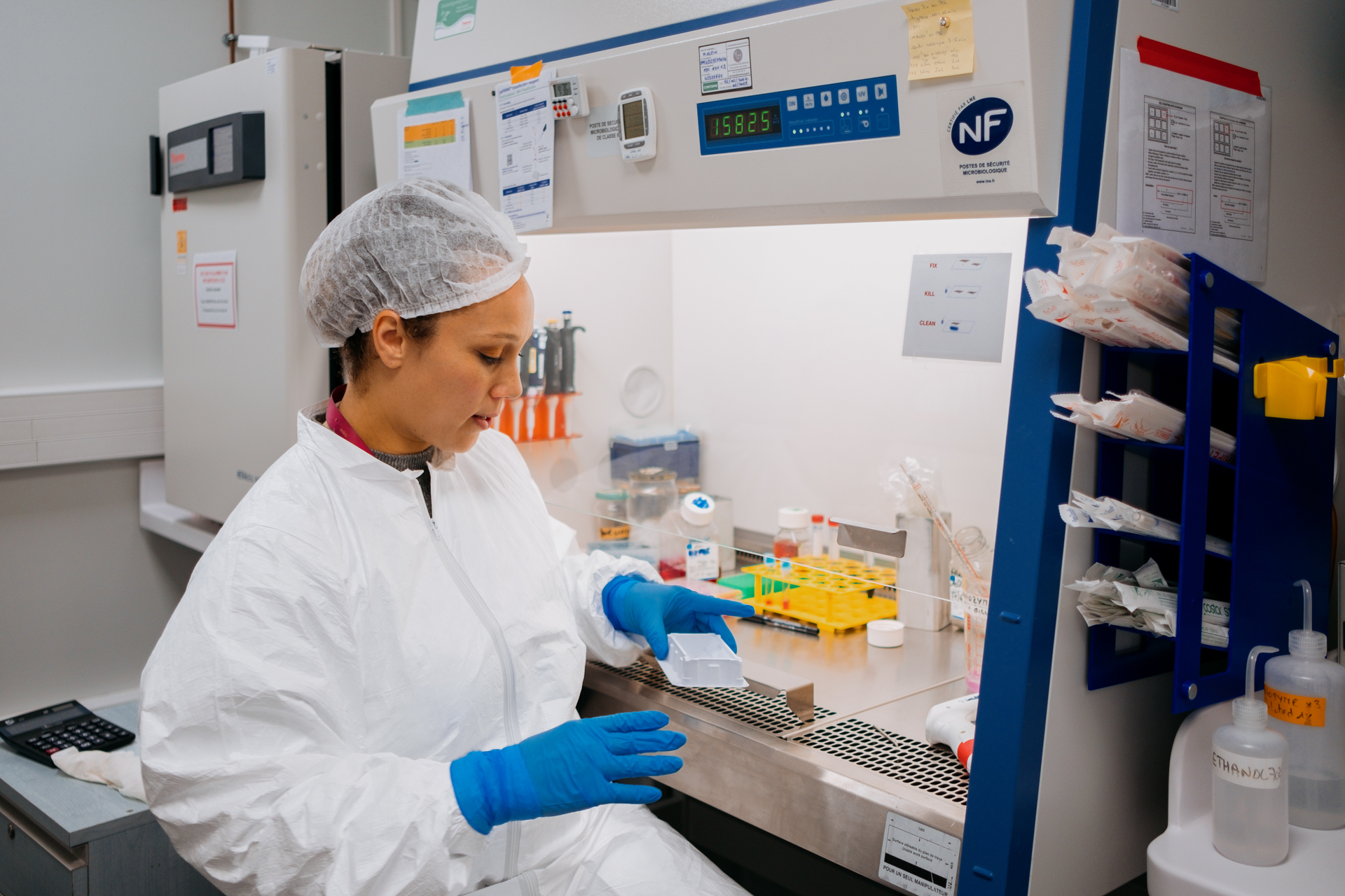 A woman in a lab coat, with her hair in a net and her hands in blue gloves, holds a sample in a lab.