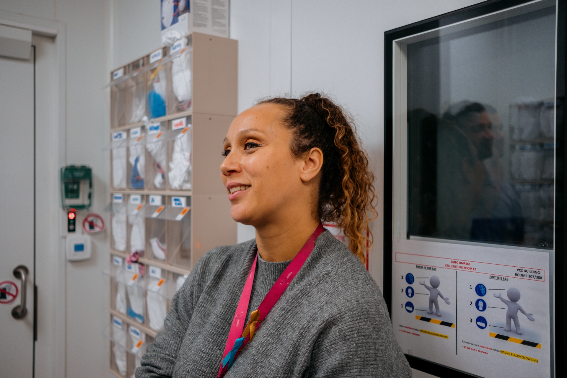 A woman in a gray sweater, with a pink lanyard, in a room with lab supplies.