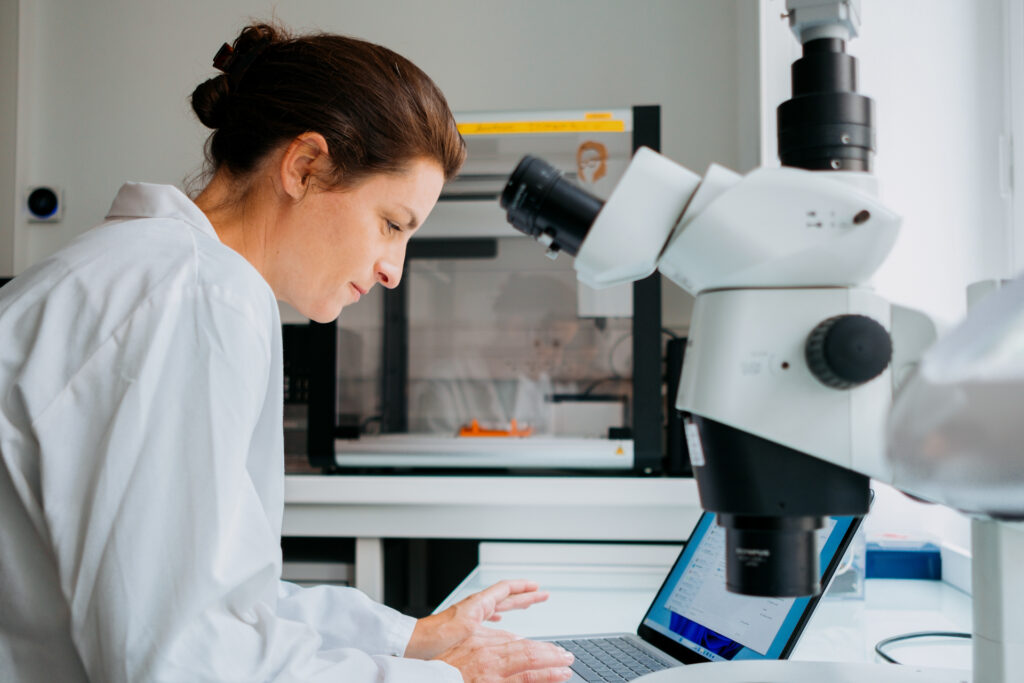 A woman with her hair pulled back, wearing a white lab coat, works on a laptop in a laboratory, with a microscope in the foreground.