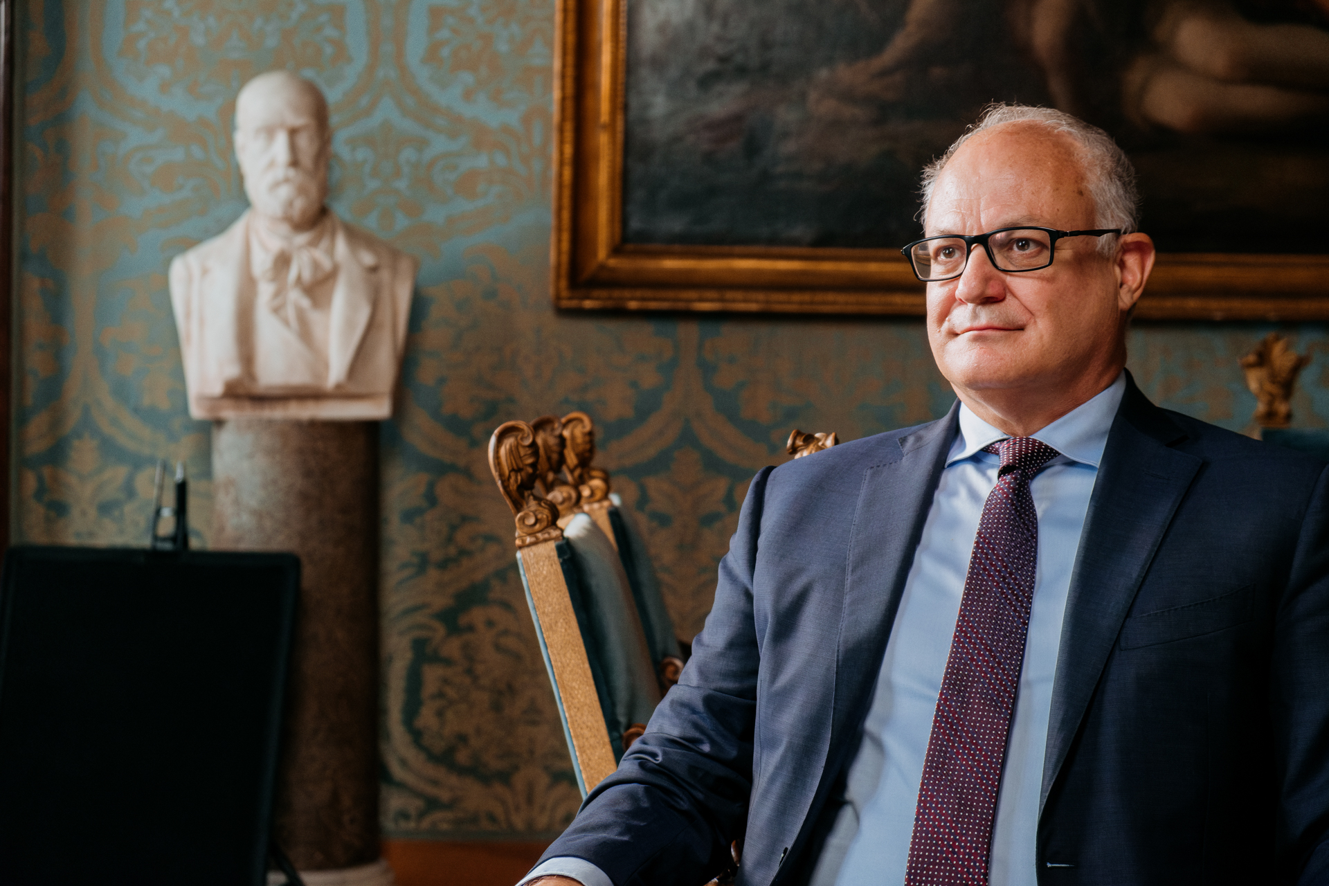 A white-haired man in a suit with dark glasses sitting in an ornate chair with a sculptural bust and painting behind him, both out of focus.