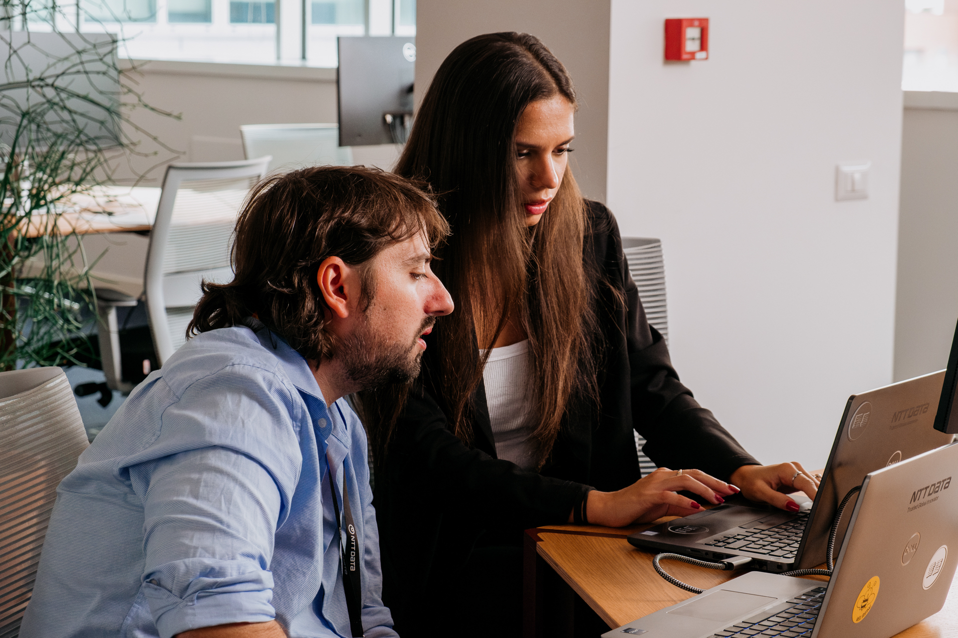 A brown-haired, bearded man in a blue shirt and a woman in a business suit with long brown hair sitting in front of a computer on a desk.