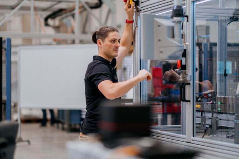 A man with a ponytail in a dark blue polo shirt working on the exterior of a machine in a glass box.