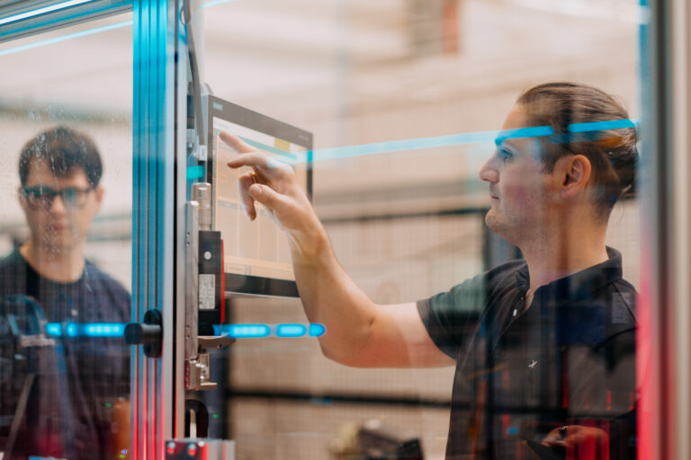 Durch reflektierendes Glas gesehen, berührt ein Mann rechts einen Computerschirm, während ein anderer die Bewegungen in der Maschine beobachtet.