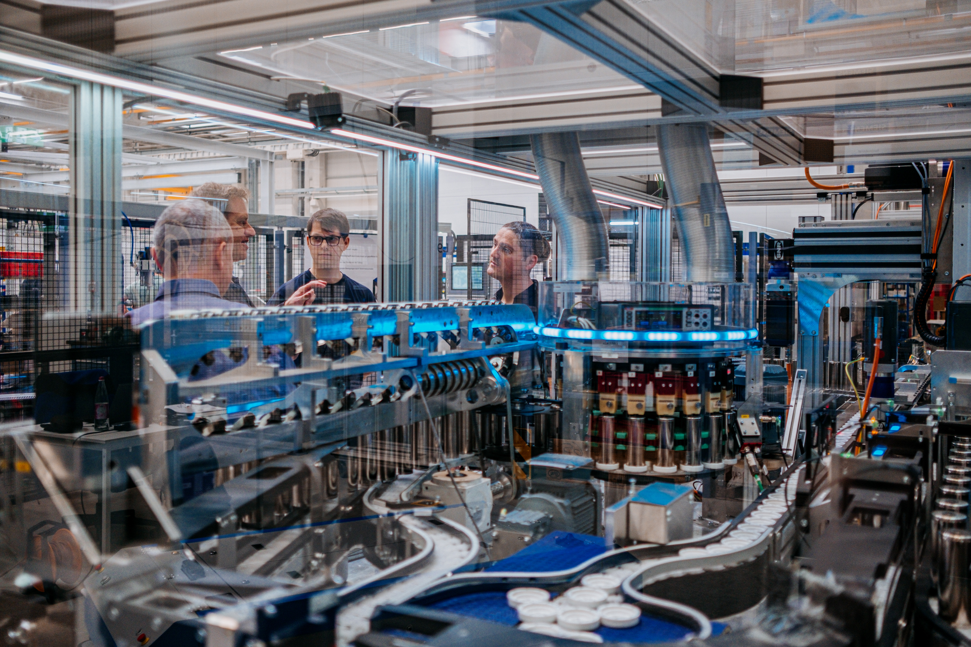 Two men with backs to camera and two men facing camera as seen through the glass protecting a complex large machine.  