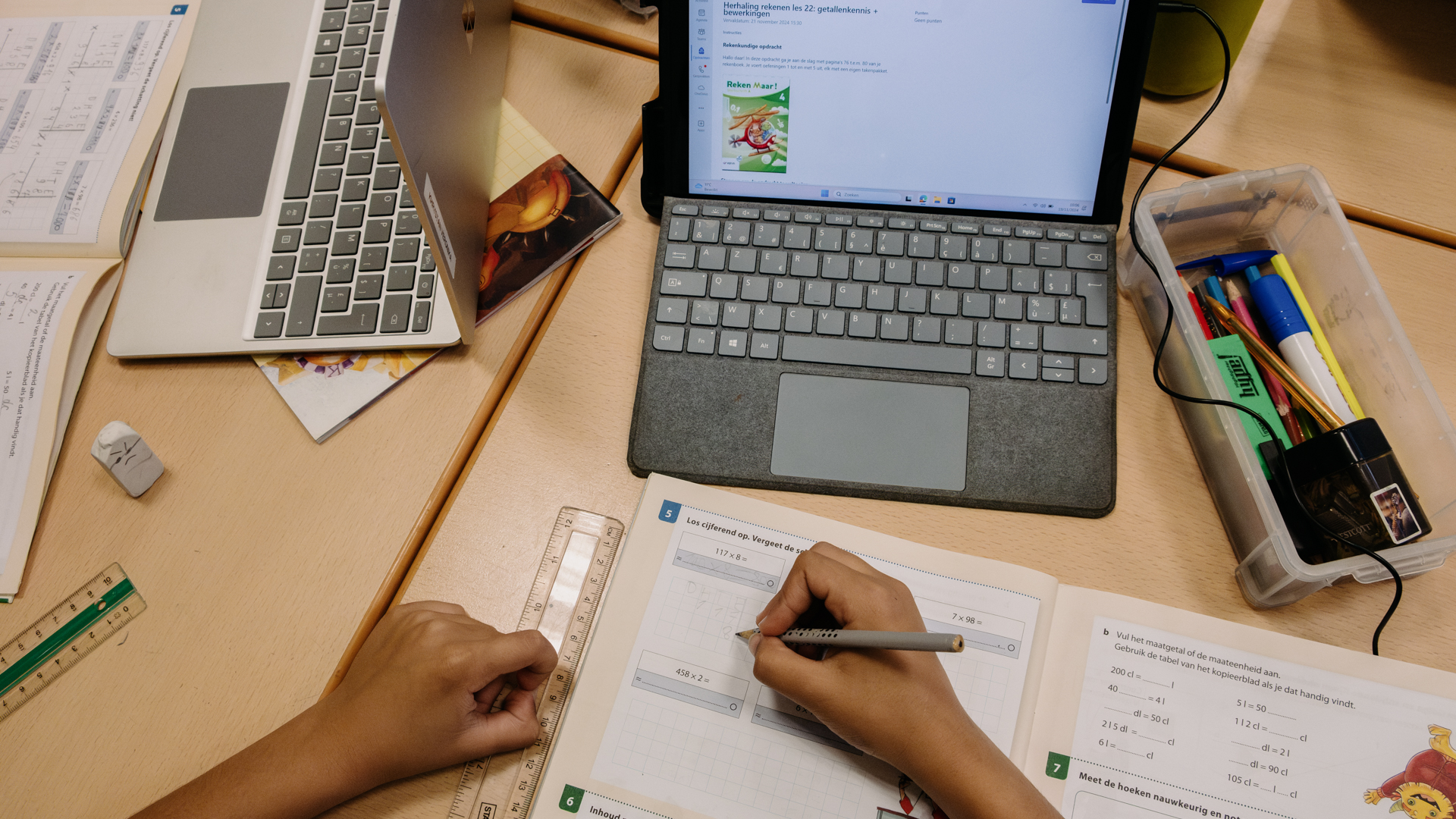 An overhead shot of a student’s arms. The right hand holds a pencil and is writing in a math workbook. A computer keyboard can be seen at the top of the image.