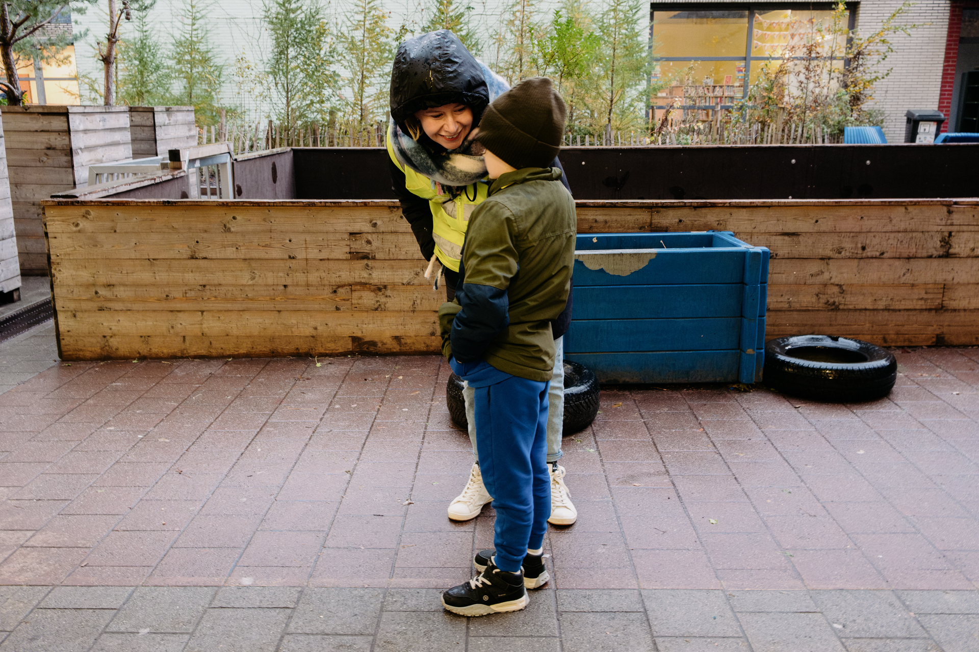 A woman in a hooded jacket leans forward to talk with a child who is also dressed for cold, wet weather. 