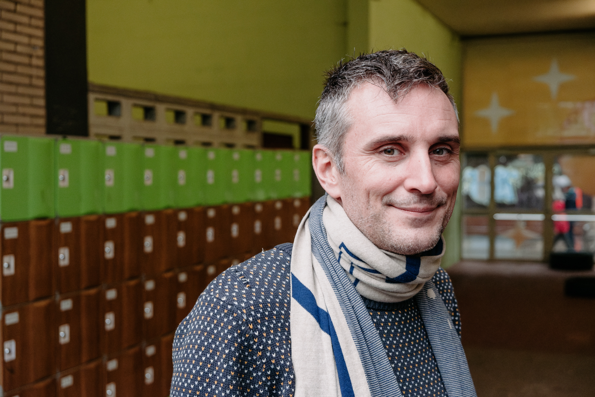 A head-and-shoulders portrait of a man with short gray hair wearing a sweater and scarf. 