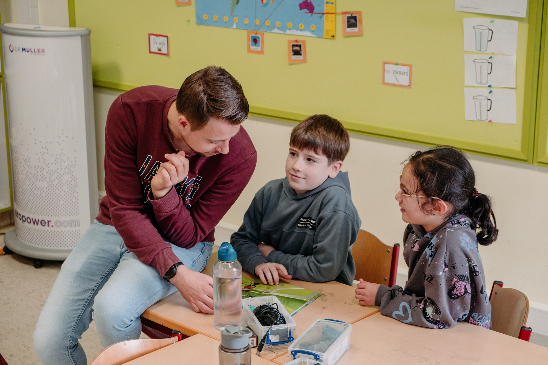 A light-haired man in a sweatshirt on the left side of the photo perches on the desk of two students, a boy, center, and a girl with a ponytail, right. 