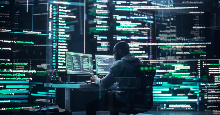 A man sits at his desk surrounded by screens that show lines of computer code.