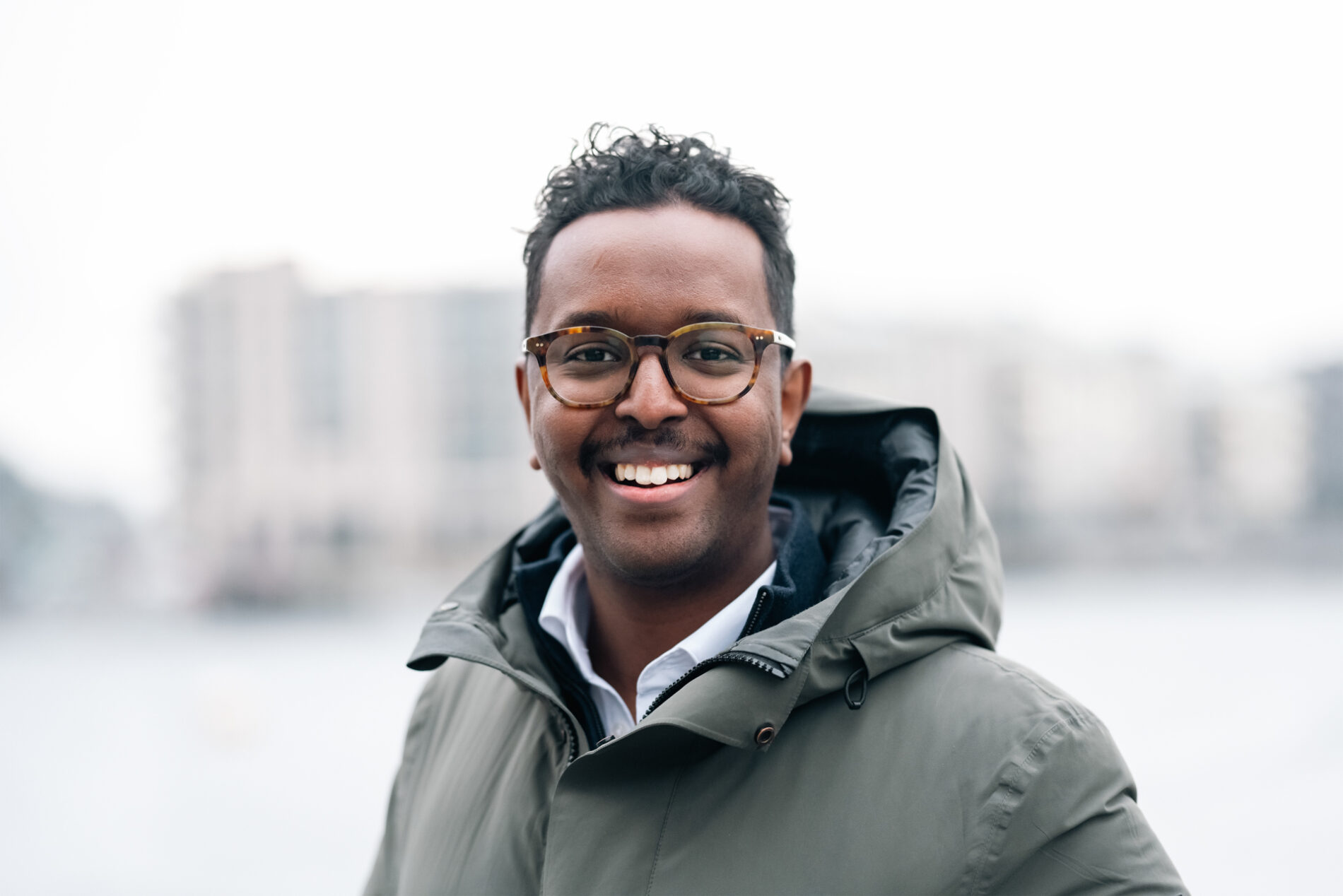 A smiling man wearing glasses and a hooded parka looks directly at the camera in this head-and-shoulders image.