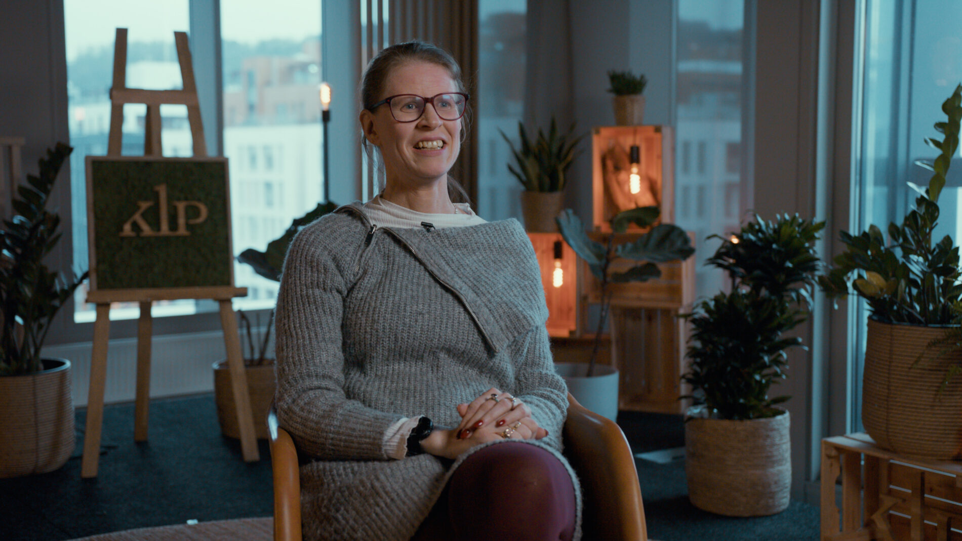 A woman with glasses seated in a modern, well-lit room with large windows and potted plants.