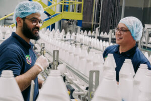 Two men in blue hairnets speaking across a line of white plastic bottles on a conveyor belt.