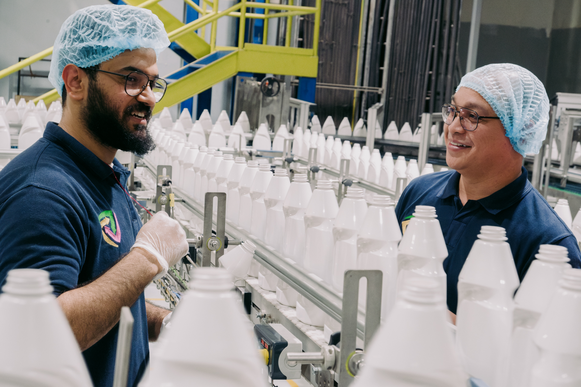 Two men in blue hairnets speaking across a line of white plastic bottles on a conveyor belt.