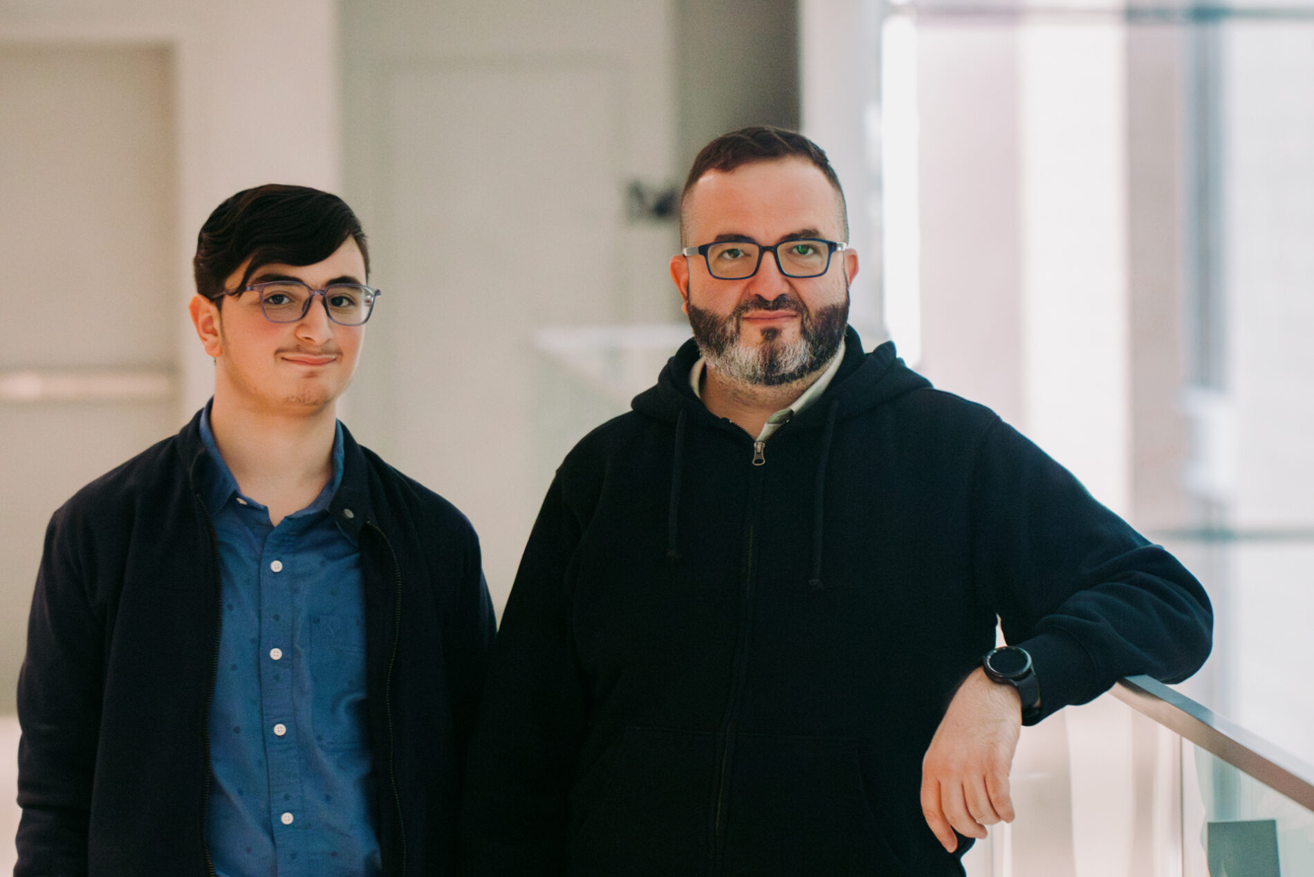 A portrait of a father, on the right with a dark sweater and glasses and a graying beard, and son, left, with glasses, a jacket and a blue buttoned shirt.