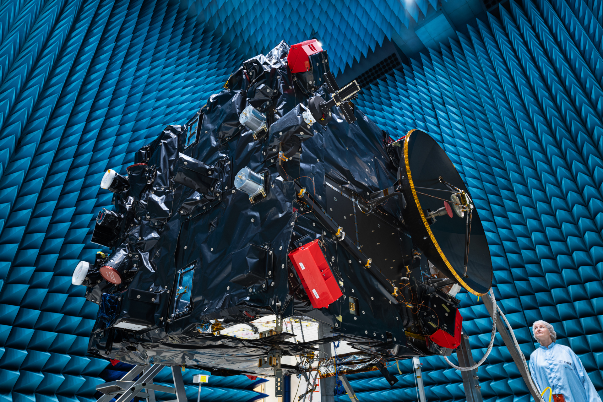 A large rectangular structure with many attachments and complex parts suspended in an enormous hangar; a woman in a sterile robe and hair cover stands on the ground at lower right.