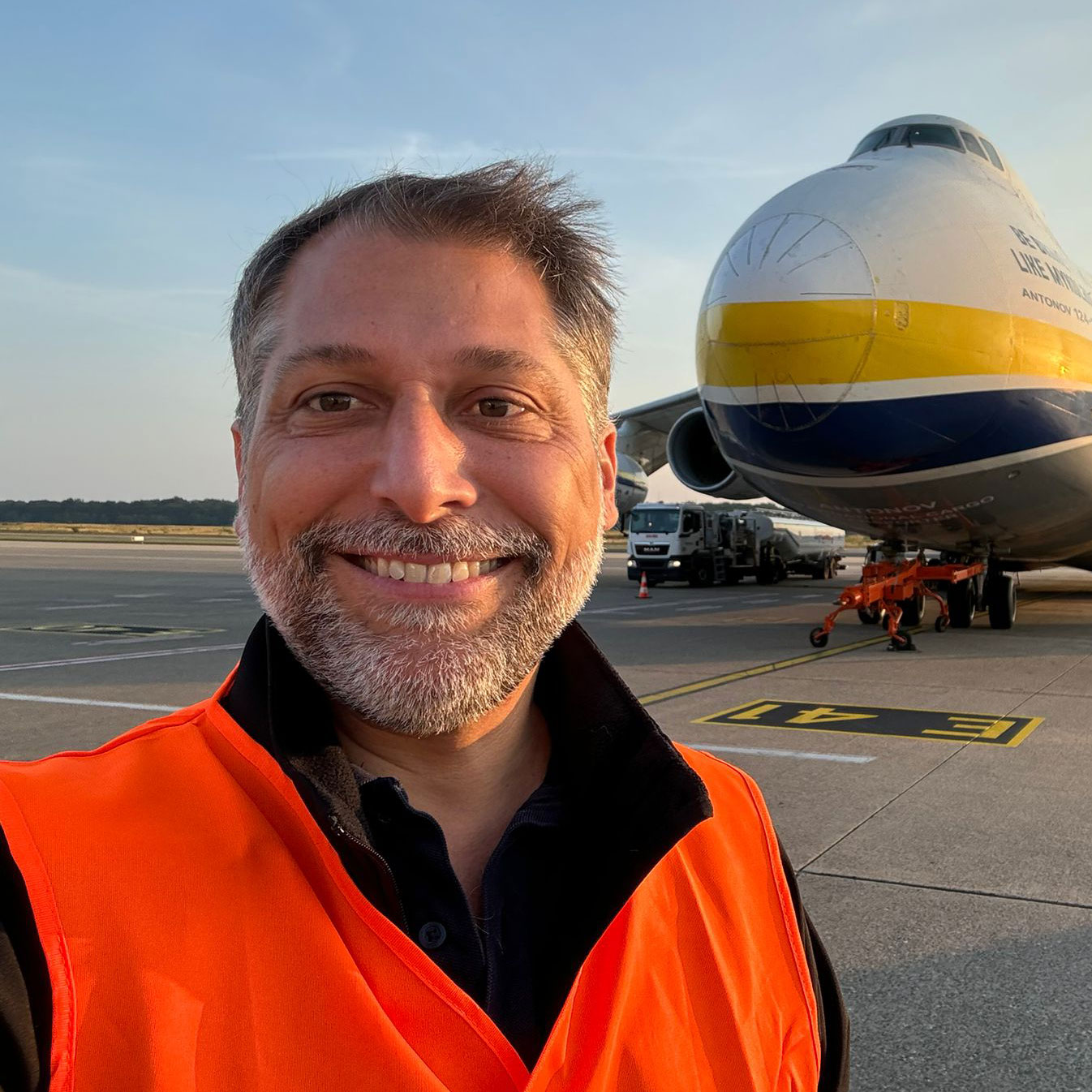 Smiling bearded man in an orange safety vest with a large jet airplane on the runway behind him.